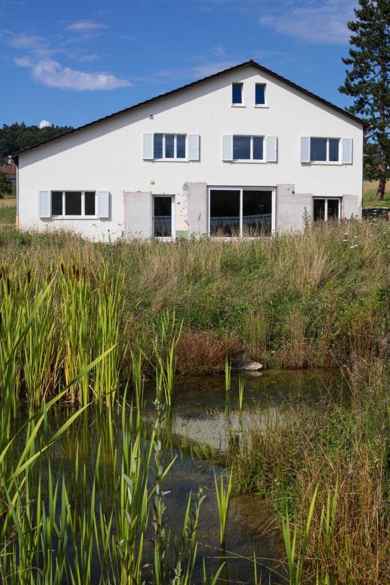 Ein weißes zweistöckiges Haus mit geschlossenen Fensterläden steht hinter hohem Gras und einem kleinen Teich, unter einem blauen Himmel mit vereinzelten Wolken - ideal für die Verwaltung oder Vermietung von Gewerbeliegenschaften.