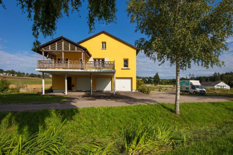 Ein leuchtend gelbes zweistöckiges Haus mit einem großen Balkon und drei Garagentoren steht an einer Landstraße, ideal für die Verwaltung oder Vermietung von Gewerbeliegenschaften. Ein weißer Lieferwagen parkt in der Nähe unter einem blauen Himmel mit vereinzelten Wolken.