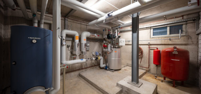 Ein sauberer, heller Heizungskeller für Gewerbeliegenschaften mit großen weißen Rohren, einem blauen Tank, einem roten Drucktank und verschiedenen Komponenten der Heizungsanlage an Betonwänden und -böden. Ein kleines Fenster lässt natürliches Licht herein.