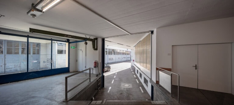 Eine Rampe führt von einem überdachten Parkplatz zu einer Gasse im Freien mit weiß gestrichelten Linien. Große Fenster säumen die linke Wand und zeigen helle Lebensräume. Auf der rechten Seite befindet sich eine geschlossene weiße Tür, während durch den offenen Eingang natürliches Licht einströmt.