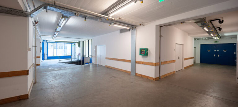 Ein sauberer, leerer Flur in einem Industriegebäude mit Betonböden, weißen Wänden und freiliegenden Lampen. Ein grüner Notfall-Defibrillator ist an der Wand angebracht - perfekt für Vermietung oder Mietangebote von modernen Gewerbeliegenschaften.