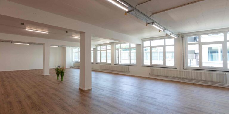 Ein geräumiger, leerer Raum mit großen Fenstern, weißen Wänden und hellem Holzfußboden - ideal für Lebensräume. Zwei Vasen mit Blumen stehen neben einer Säule, während natürliches Licht dieses einladende Mietangebot erfüllt.