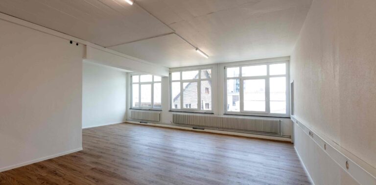 Ein heller, leerer Raum mit großen Fenstern, weißen Wänden und Holzfußböden - ideale Lebensräume. Sonnenlicht strömt herein, Heizkörper sitzen unter den Fenstern, und draußen ist ein Gebäude mit schrägem Dach zu sehen - eine gute Wahl unter den Mietangeboten.