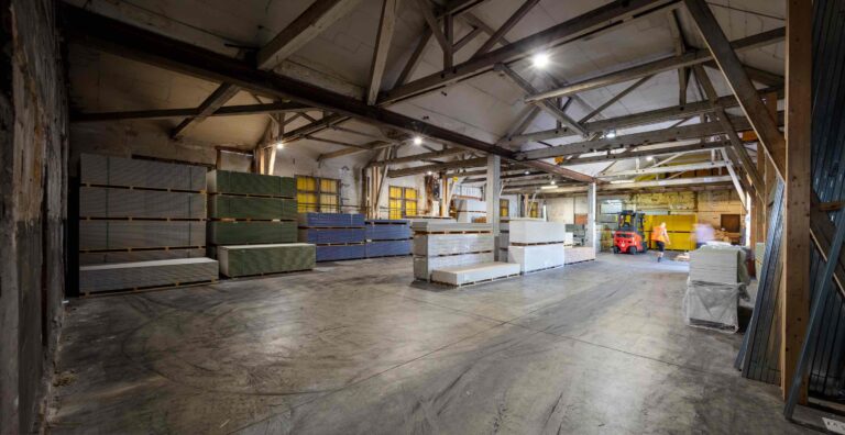 Eine geräumige Lagerhalle mit sichtbaren Balken, Betonböden und Stapeln von Trockenbauplatten in verschiedenen Farben, die fein säuberlich an den Wänden angeordnet sind. Helle Oberlichter erhellen dieses ideale Gewerbeliegenschaften Mietangebot.