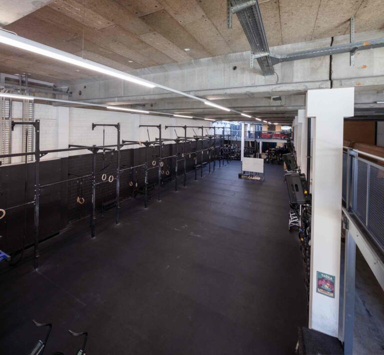 Eine geräumige, leere Turnhalle mit schwarzem Gummibodenbelag, mehreren Klimmzugstangen, Gymnastikringen und Fitnessgeräten an den Wänden unter einer Betondecke mit freiliegenden Rohren - ideal für die Vermietung von attraktiven Gewerbeliegenschaften.