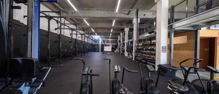 Ein geräumiges, modernes Fitnessstudio mit schwarzem Bodenbelag, Reihen von stationären Fahrrädern im Vordergrund und Hantelständern, Klimmzugständern und Fitnessgeräten an den Wänden - perfekte Lebensräume für Fitness. Sichtbetondecke und Oberlichter.