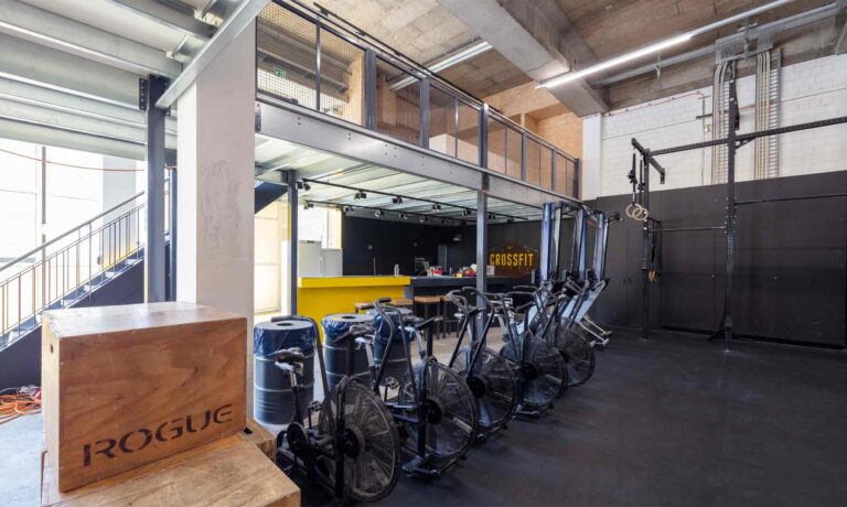 Ein modernes CrossFit-Fitnessstudio mit stationären Fahrrädern, hölzernen plyometrischen Boxen und einem gelben Empfangstresen unter einem Zwischengeschoss - dieser Industrieraum von Lebensräume bietet einzigartige funktionelle Fitness und modernes Design.