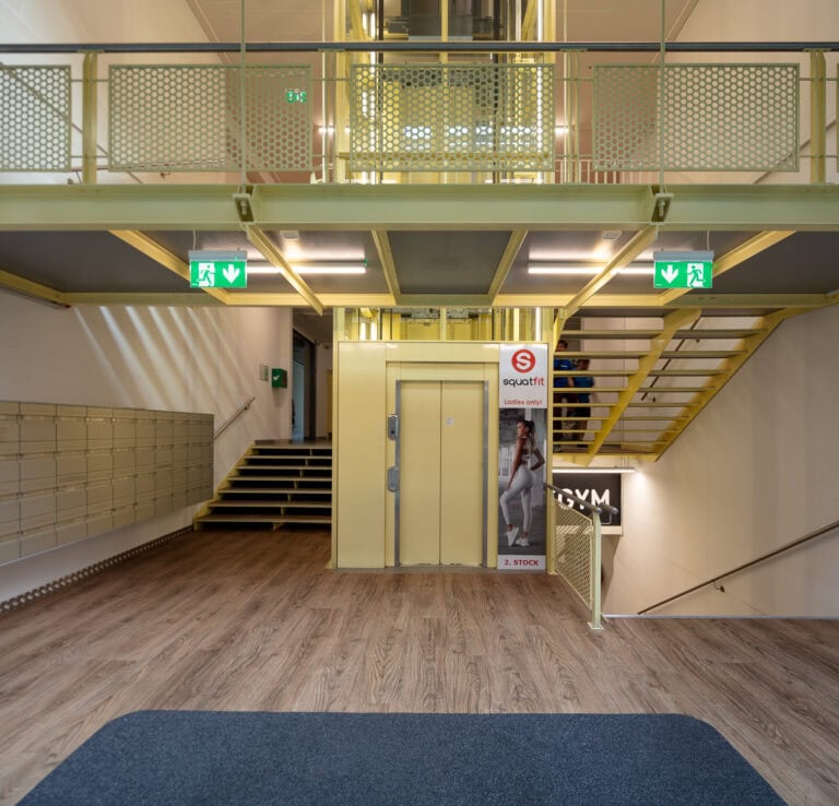 Ein modernes Foyer am Leimgrubenweg 9 in Basel hat einen Holzboden, ein gelbes Geländer, eine Treppe, die nach oben und unten führt, Ausgangsschilder, Briefkästen an der linken Wand und ein teilweise sichtbares Schild eines Fitnessstudios im Erdgeschoss.