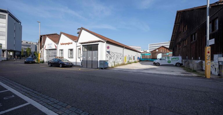 Eine Straße mit geparkten Autos neben Gebäuden, darunter eine weiße Lagerhalle mit Graffiti, eine Holzkonstruktion und ein weißer Lieferwagen mit der Aufschrift 