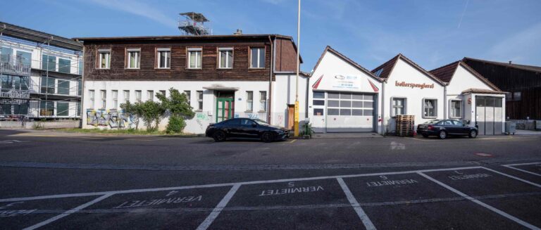 Eine Reihe von Gewerbeliegenschaften, darunter ein zweistöckiges Holzgebäude und eine weiße Autowerkstatt mit drei Garagentoren. Zwei geparkte Autos und leere Parkplätze mit der Aufschrift 