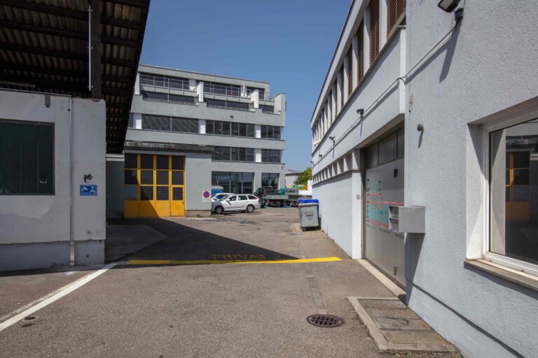 Ein gepflasterter Gewerbehof mit grauen Gebäuden, einem gelben Garagentor, geparkten Autos und einer 