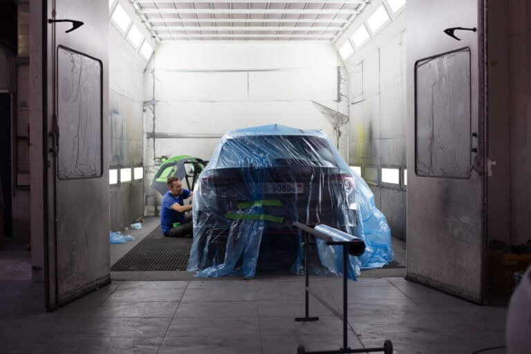 Eine Person in einem blauen Hemd arbeitet an einem Auto, das in einer Karosseriewerkstatt mit Plastikfolien abgedeckt ist, wo helle Lichter über dem Kopf und offene Türen dynamische Lebensräume und aktive Gewerbeliegenschaften hervorheben.