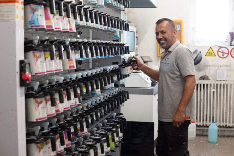 Ein lächelnder Mann in einem grauen Polohemd steht neben Regalen voller bunter Farbdosen und hält eine Dose in der Hand. Der helle Raum ist mit industriellen Geräten und Warnschildern ausgestattet - ideal für alle, die auf der Suche nach Gewerbeliegenschaften oder einzigartigen Lebensräumen sind.