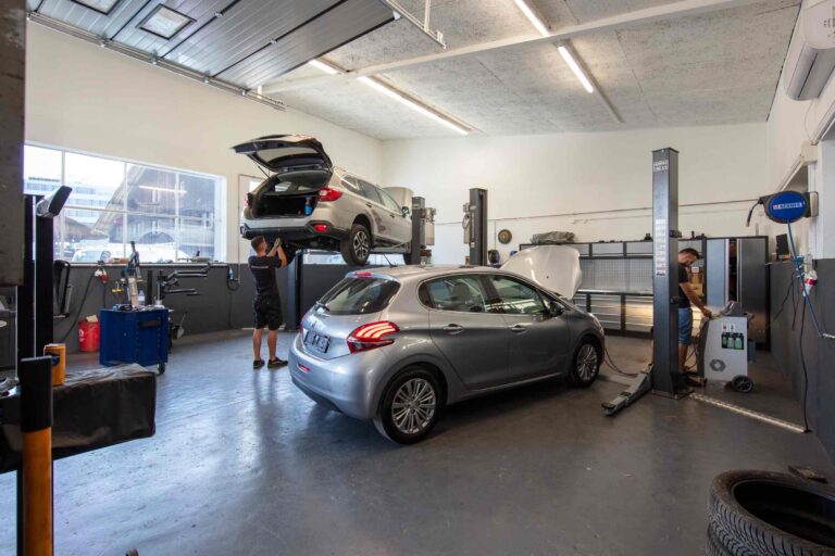 Ein Mechaniker arbeitet an einem silbernen Geländewagen, der auf einer Hebebühne in einer hellen, gut organisierten Autowerkstatt mit Mietangeboten angehoben wird, während eine andere Person an der Motorhaube eines geparkten silbernen Schrägheckwagens arbeitet. Werkzeuge und Ausrüstung sind rund um die Werkstatt zu sehen.