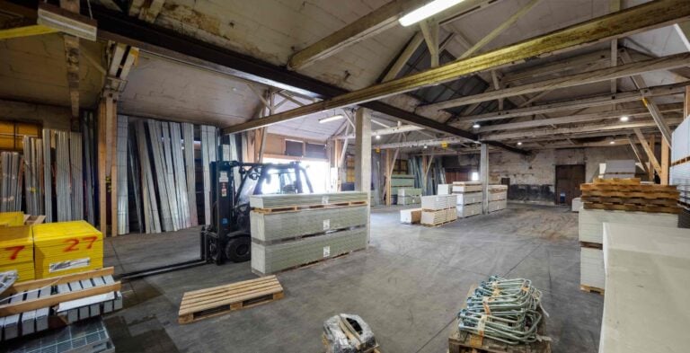 Ein geräumiges Lagerhaus, ideal für die Vermietung von Gewerbeliegenschaften, mit einem Gabelstapler, der Material bewegt, Stapeln von Baumaterialien, Holzpaletten und Metallträgern unter freiliegenden Holzdeckenbalken und Leuchtstoffröhrenbeleuchtung.