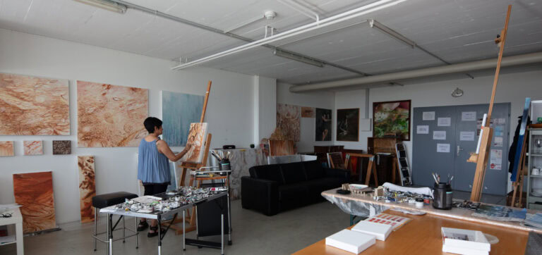 Eine Person steht an einer Staffelei und malt in einem hellen, geräumigen Kunstatelier - einem der inspirierenden Lebensräume in einzigartigen Gewerbeliegenschaften. Große Gemälde schmücken die Wände, die Tische sind voll mit Material, und durch die Fenster strömt natürliches Licht.