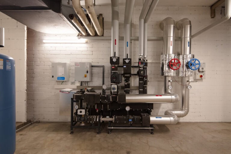 Industrieller Maschinenraum in Gewerbeliegenschaften mit freiliegenden Rohren, Ventilen, Messgeräten und Schalttafeln an einer weißen Backsteinwand. Blaue und rote Radventile heben sich von zwei großen silbernen Rohren ab; andere Messgeräte weisen auf effiziente Vermietungssysteme hin.