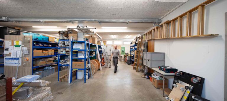 Eine Person geht durch eine Lagerwerkstatt mit Regalen mit Kisten, Werkzeugen und Materialien auf beiden Seiten. Der gut beleuchtete Raum mit Betonboden zeigt organisierte Lagerbereiche, ideal für die Verwaltung moderner Gewerbeliegenschaften.