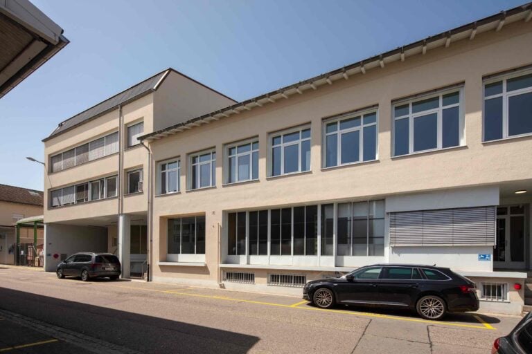 Ein modernes beigefarbenes Gebäude mit großen rechteckigen Fenstern und einem schrägen Dach bietet attraktive Mietangebote. An einem sonnigen Tag sind mehrere Autos auf der Straße davor geparkt.