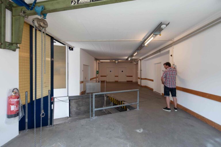 Ein Mann in einem karierten Hemd steht an einer Wand in einer Industriehalle mit weißen Wänden, Deckenbeleuchtung und einer Rampe, die gut geführte Gewerbeliegenschaften zur Aufwertung moderner Lebensräume zeigt. An der linken Wand ist ein Feuerlöscher angebracht.