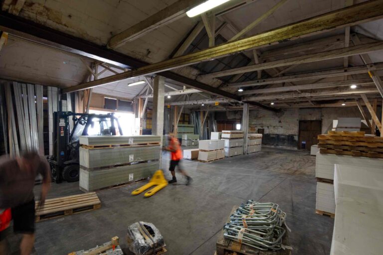 Ein Lagerhaus mit gestapelten Paletten von Baumaterialien, einem Gabelstapler und zwei Arbeitern in Bewegung - ideal für Vermietung oder Verwaltung. Der Raum ist mit freiliegenden Balken und Industriebeleuchtung ausgestattet.