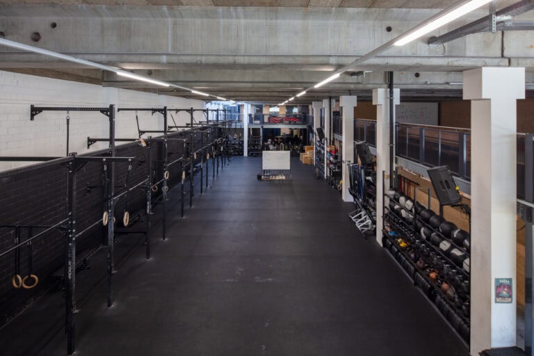 Ein geräumiger, moderner Fitnessraum mit schwarzem Gummiboden und Wandregalen bietet vielseitige Lebensräume mit Gymnastikringen, freien Gewichten, Medizinbällen und anderen Fitnessgeräten, die ordentlich unter einer Betondecke angeordnet sind.