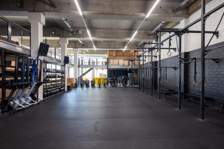 Ein geräumiger, moderner Fitnessraum mit schwarzem Gummiboden, Hantelständern, Rudergeräten und Gymnastikringen bietet helle Lebensräume; große Fenster und industrielle Beleuchtung bereichern diesen offenen Trainingsbereich, der in unseren Mietangeboten vorgestellt wird.