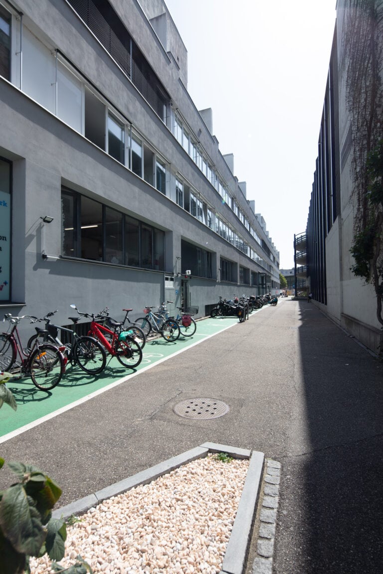 Eine schmale Gasse zwischen zwei modernen Gebäuden, die von der Verwaltung verwaltet werden, mit Fahrrädern, die auf der linken Seite auf einem grünen Radweg abgestellt sind, und einem Fleck mit weißen Steinen im Vordergrund unter einem sonnigen Himmel.