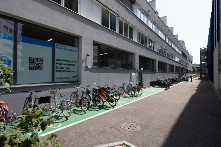 Eine Reihe von Fahrrädern ist an einem sonnigen Tag auf einem grünen Radweg neben modernen Gewerbeliegenschaften mit großen Fenstern entlang einer schmalen Straße geparkt. Ein großes Fenster zeigt Text und einen QR-Code, der zu Vermietungsanfragen einlädt.