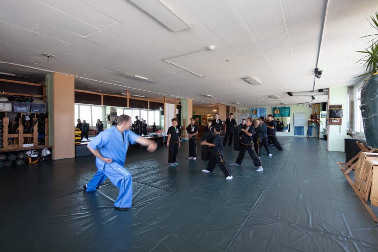 Ein Kampfsportlehrer in Blau leitet eine Gruppe von Schülern in schwarzen Uniformen an, die in einem geräumigen, gut beleuchteten Studio mit Matten auf dem Boden gemeinsam Bewegungen üben - ein dynamischer Lebensraum, perfekt für das Training.