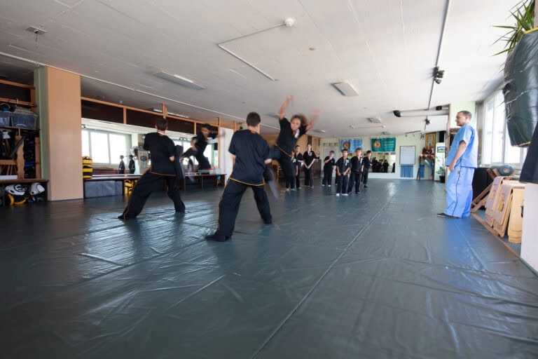 Eine Gruppe von Kindern in Kampfsportuniformen übt in einem geräumigen, hell erleuchteten Studio, das für die Vermietung zur Verfügung steht, Bewegungen, während ein Ausbilder in Blau von der Seite zuschaut. Einige Schüler befinden sich mitten in der Aktion und demonstrieren dynamische Techniken.