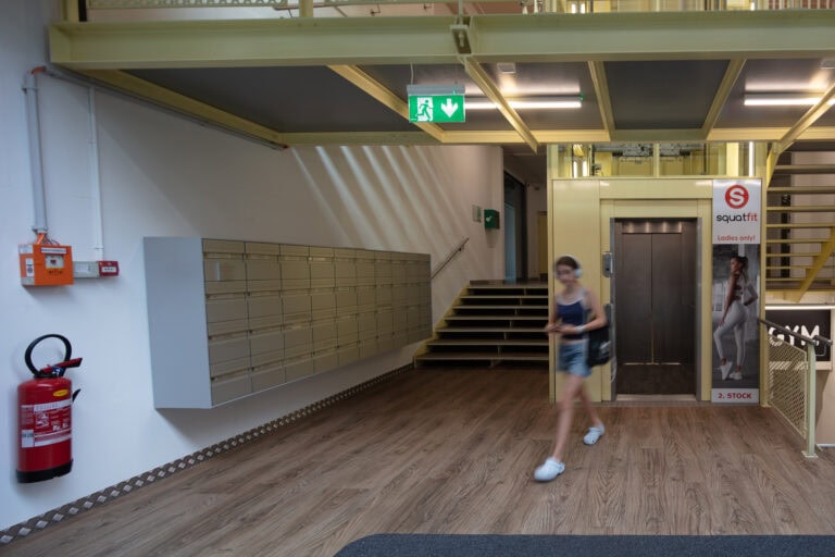 Eine Frau geht durch ein modernes Foyer mit Briefkästen, einem Feuerlöscher, einer Treppe, einem Aufzug und einem Schild mit der Aufschrift 