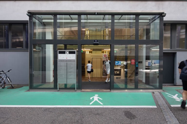 Ein gläserner Eingang zu einem modernen Gebäude mit zwei Personen in der Nähe des Eingangs. Ein grüner Weg, der mit einem Fußgängersymbol gekennzeichnet ist, führt zum Eingang dieser attraktiven Gewerbeliegenschaften. Links sind Fahrräder geparkt, rechts geht eine weitere Person.