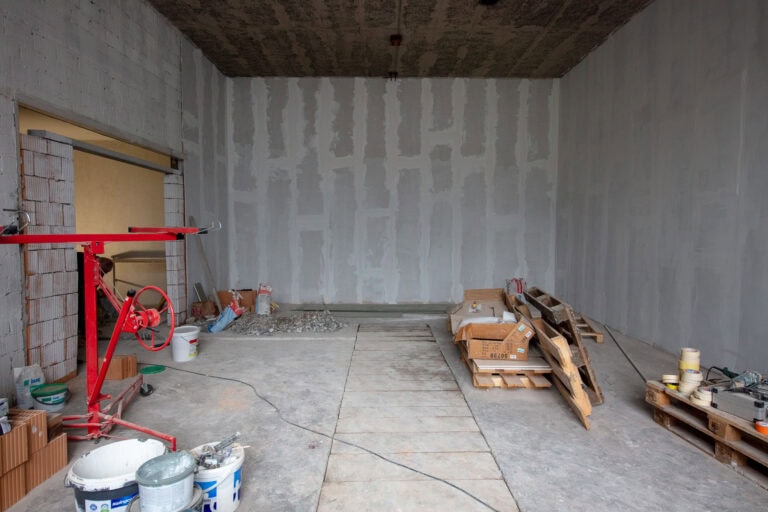 Ein unfertiger Lebensräume-Raum mit grauer Trockenbauwand, Baumaterialien, Werkzeugen, Holzpaletten und Eimern, die auf dem Betonboden verstreut sind. Die Decke ist freigelegt, die Wände sind teilweise gestrichen und geflickt.