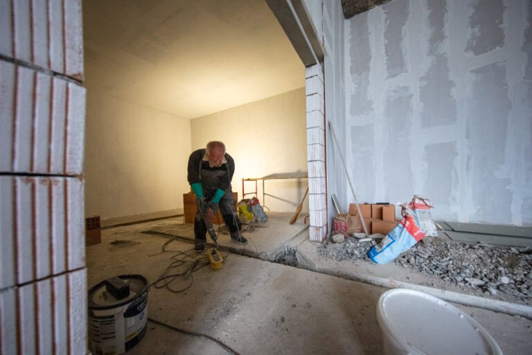 Eine Person in Arbeitskleidung bückt sich, während sie mit Bauwerkzeugen in einem unfertigen Raum mit freiliegenden Wänden, verstreuten Materialien und Geräten auf dem Boden arbeitet - neue Lebensräume für Mietangebote gestalten.