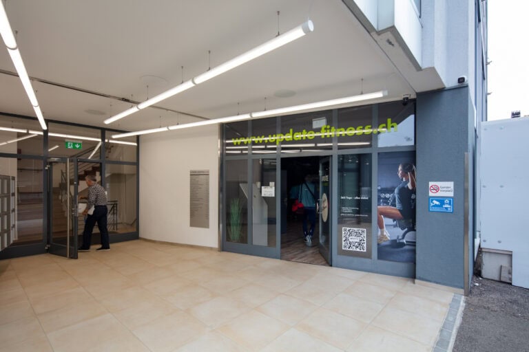 Ein moderner Gebäudeeingang mit Glastüren; ein Mann geht hinein. Neben den Türen bieten Gewerbeliegenschaften ein Fitnessstudio mit großen Fenstern, einer Website-URL darüber, einem Poster mit einer trainierenden Frau und einem sichtbaren QR-Code.