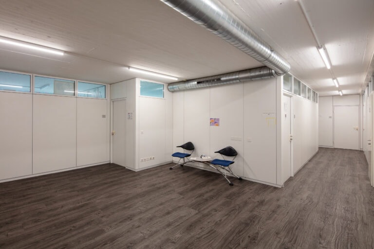 Ein sauberer, moderner Flur mit weißen Wänden, holzgemustertem Boden, Deckenleuchten, zwei blauen Stühlen und einem kleinen Tisch an der Wand. Große Lüftungskanäle an der Decke verleihen diesem Gewerbeliegenschaften-Raum einen modernen Touch.