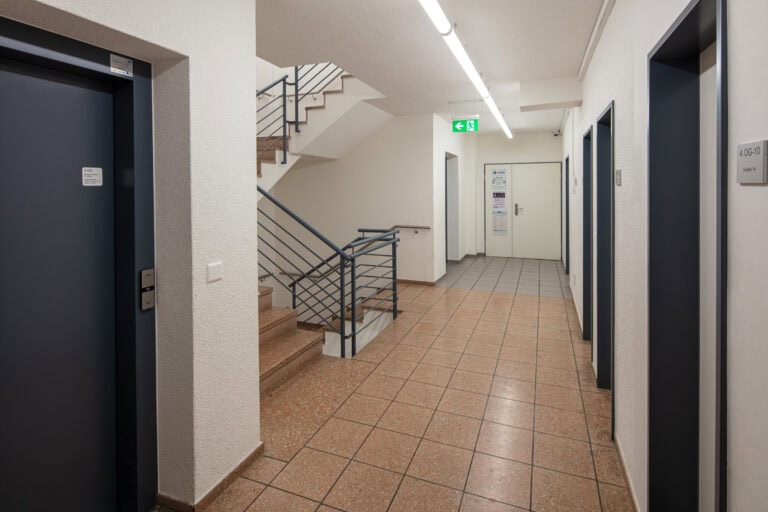 Ein sauberer Flur in einem Gebäude mit gefliesten Böden, zwei Aufzugstüren auf einer Seite, eine Treppe mit Metallgeländer - ideal für Gewerbeliegenschaften -, Leuchtstofflampen, ein gefliester Treppenabsatz und ein Ausgangsschild über einer Tür am Ende.