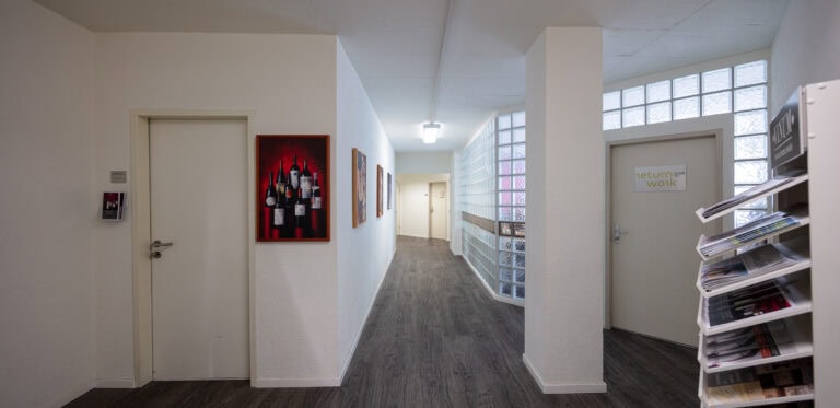 In einem Flur mit weißen Wänden und dunklem Holzfußboden befinden sich geschlossene Türen, Wandkunst, ein Regal mit Mietangeboten, Glasbausteinfenster und eine Tür mit der Aufschrift 