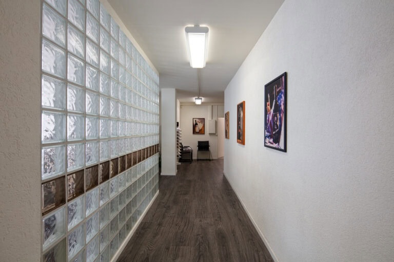 Ein Flur mit Holzfußboden, einer Wand aus Glasbausteinen auf der linken Seite und einer weißen Wand mit bunter Kunst auf der rechten Seite. Leuchtstoffröhren an der Decke erhellen diese einladenden Lebensräume und führen zu einem Bürobereich mit Regalen und einem Stuhl am Ende.