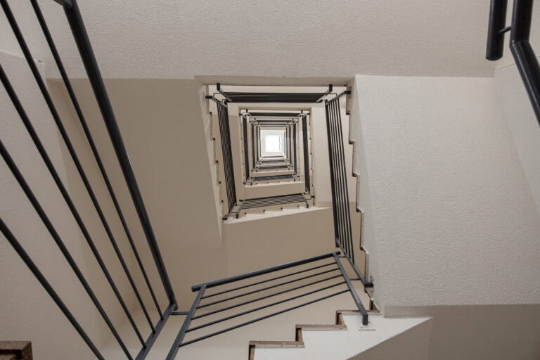 Ein Blick auf eine quadratische, mehrstöckige Treppe mit Metallgeländern und weißen Wänden, die einen geometrischen, tunnelartigen Effekt erzeugen - perfekt für moderne Lebensräume oder Gewerbeliegenschaften - und oben in eine helle Öffnung münden.