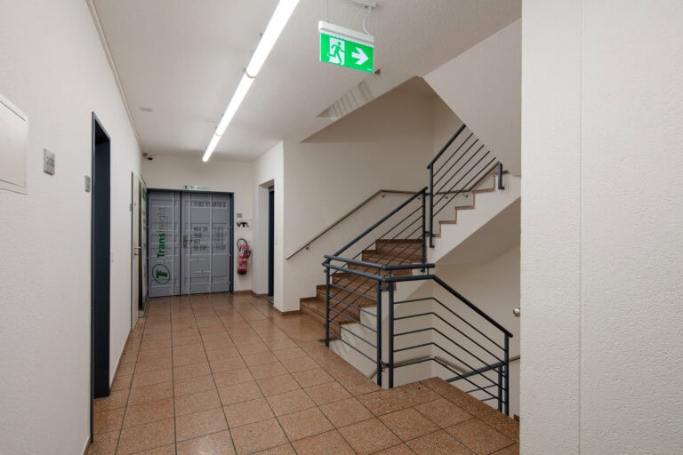 Ein sauberer, gefliester Flur in einem Gebäude mit einer Treppe, Metallgeländern und einem beleuchteten grünen Ausgangsschild an der Decke - ideale Lebensräume. Am Ende des Flurs stehen graue Spinde und ein Feuerlöscher an der Wand.