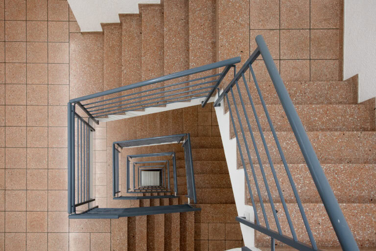 Draufsicht auf eine Wendeltreppe mit braun gefliesten Stufen und blaugrauen Metallgeländern, die beim Abstieg ein geometrisches Muster bilden - ein elegantes Merkmal, das häufig in modernen Verwaltungsgebäuden und kuratierten Lebensräumen zu finden ist.