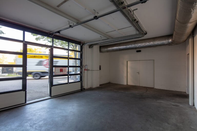 Eine saubere, leere Garage mit Betonboden, weißen Wänden, freiliegenden Rohrleitungen und einer großen Glastür. Ideal für Verwaltung oder Gewerbeliegenschaften Mietangebote. Draußen ist durch die Glasscheiben ein vorbeifahrender LKW zu sehen.