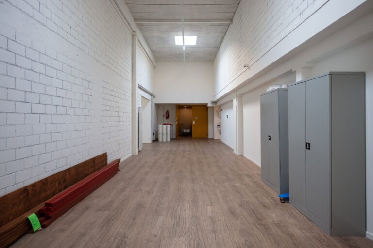 Ein breiter, leerer Flur mit hellem Holzfußboden, weißen Ziegeln und glatten Wänden, einem Oberlicht, Metallschränken auf der rechten Seite und roten Balken, die links an der Rückwand gestapelt sind - ideal für die Verwaltung oder flexible Lebensräume.