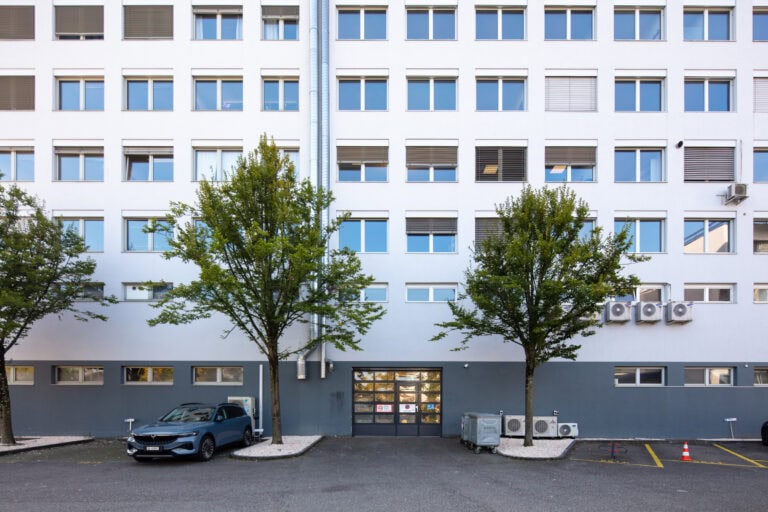 Ein geparktes Auto steht unter zwei Bäumen vor einem modernen, weißen, mehrstöckigen Verwaltungsgebäude mit Fensterreihen und Klimageräten. Die gepflasterte Fläche ist mit kleinen Steinen und Markierungen gesäumt, die einladende Lebensräume für zukünftige Mietangebote bieten.