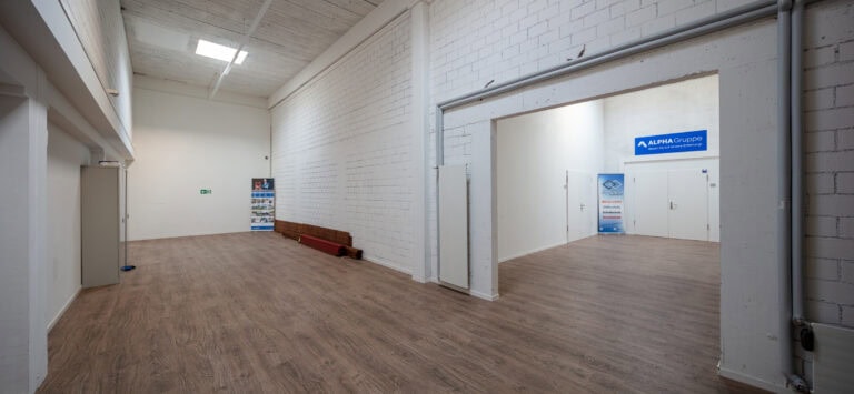 Ein geräumiger Flur mit weißen Backsteinwänden, hohen Decken, Holzböden und Leuchtstoffröhrenbeleuchtung. Ein offener Durchgang führt zu einem Verwaltungsbereich mit Firmenschildern und weißen Türen. An der gegenüberliegenden Wand ist ein Schild mit einem Notausgang zu sehen.