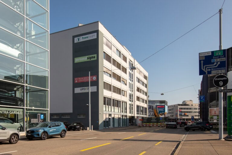 Eine Stadtstraße mit modernen Bürogebäuden, Gewerbeliegenschaften, geparkten Autos, Verkehrsschildern und einem gläsernen Parkhaus unter strahlend blauem Himmel. An den Wänden einiger Bürogebäude hängen große Geschäftsschilder.
