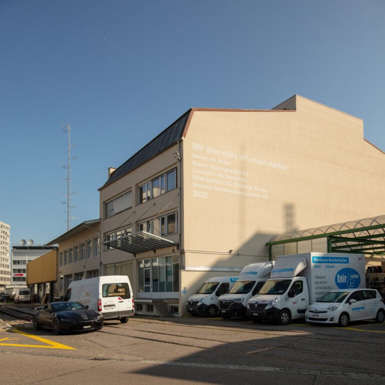 Ein beiges Industriegebäude mit schweizerdeutschem Text an der Wand, mehrere geparkte Lieferwagen mit Firmenlogos, ein schwarzes Auto und ein teilweise bewölkter blauer Himmel - ideal für Lebensräume und Mietangebote.
