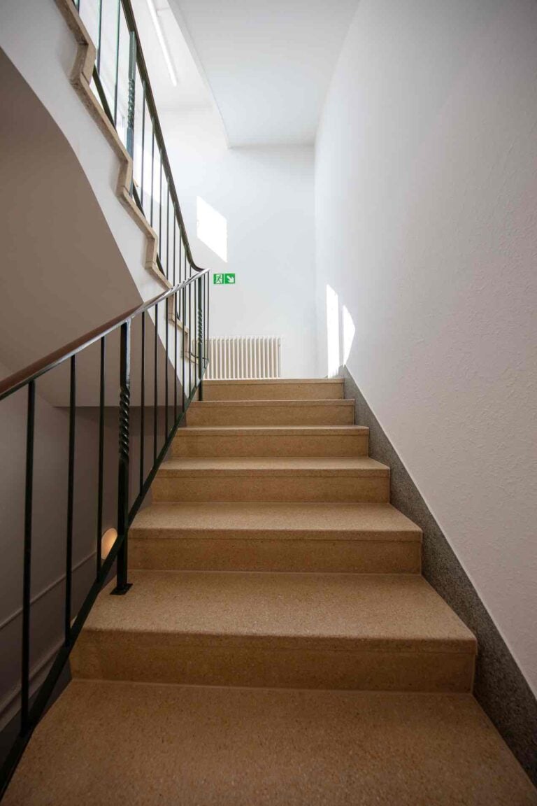 Eine Treppe mit beigen Stufen und schwarzem Handlauf führt zu einem hellen Treppenabsatz in dieser Verwaltung, wo ein weißer Heizkörper und ein Notausgangsschild zu sehen sind; durch die Fenster darüber fällt natürliches Licht ein.